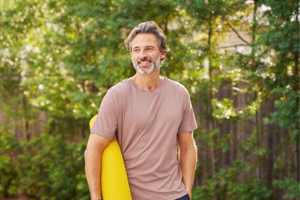 A smiling middle-aged man with graying hair and a beard stands outdoors in a backyard, holding a bright yellow yoga mat. wearing a taupe-brown t-shirt.
