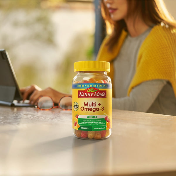 A bottle of Nature Made Multi + Omega-3 dietary supplements sits on a wooden table in a warm, sunlit room, with a woman working on a laptop in the softly blurred background.