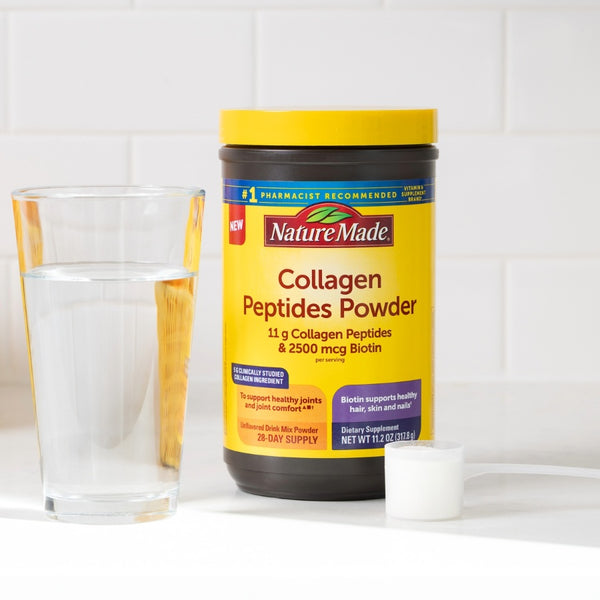 A tub of Nature Made Collagen Peptides Powder with a yellow label and cap, shown next to a glass of water and a white scoop on a white surface against a white tiled background.