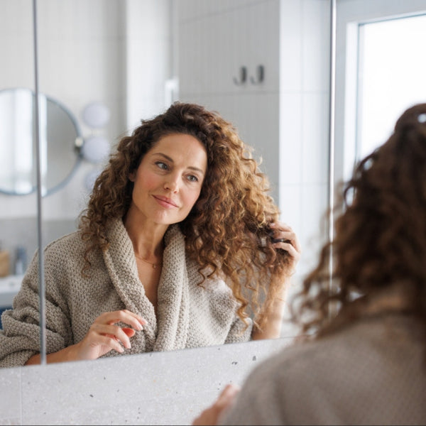 Woman in a beige robe with curly hair looking at herself in a bathroom mirror.