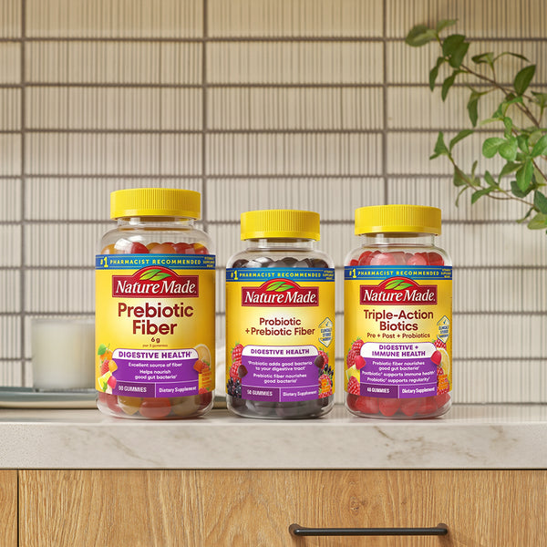 Three Nature Made supplement bottles on a kitchen counter: Prebiotic Fiber, Probiotic + Prebiotic Fiber, and Triple-Action Biotics, with a tiled wall and plant in the background.