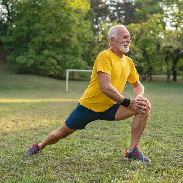 An older man with white hair and a beard is stretching on a grassy field. He is wearing a bright yellow t-shirt, dark blue athletic shorts, and gray running shoes with red accents.