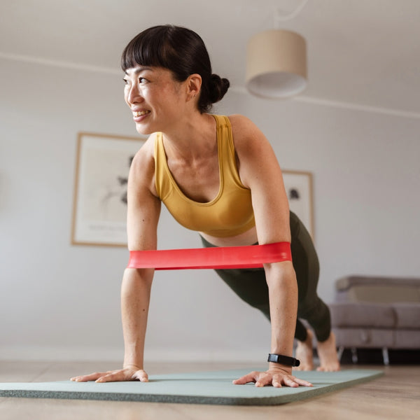 A woman in a yellow sports bra and dark leggings does a push-up on a yoga mat, with a red resistance band around her waist in a bright home setting.