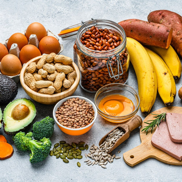 A styled flat-lay of various foods on a light gray textured surface, including eggs, peanuts, sweet potatoes, bananas, broccoli, avocado, seeds, and sliced meat, with a large glass jar of red beans prominently featured in the center.