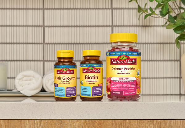 Three NatureMade supplement bottles on a countertop: Hair Growth, Biotin, and Collagen Peptides + C, with a tiled wall and rolled towels in the background.