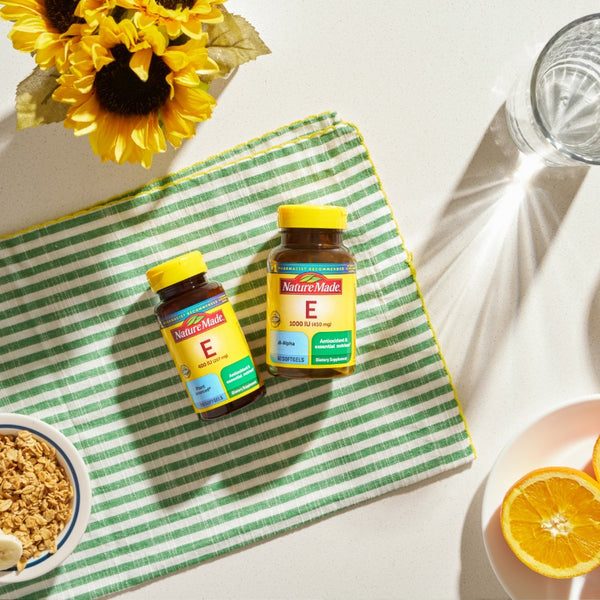 Two bottles of Nature Made Vitamin E on a green and white striped towel with sunflowers and oranges in the background.